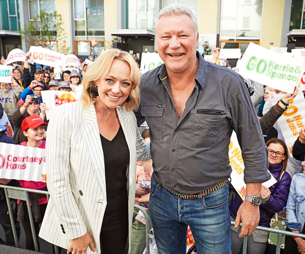 Auction day scene from "The Block" with two hosts smiling and a crowd holding supportive signs in the background.