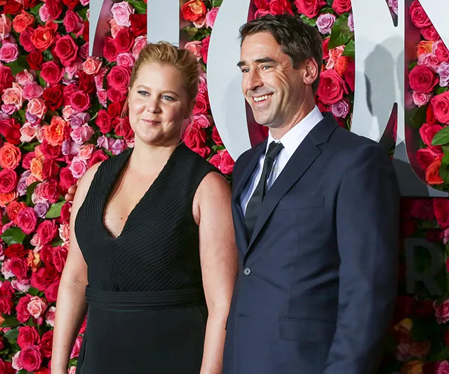 A woman in a black dress and a man in a suit stand in front of a floral wall.