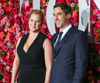 A woman in a black dress and a man in a suit stand in front of a floral wall.