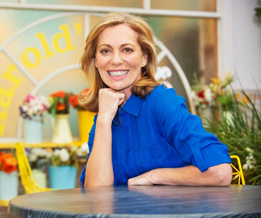 Kerry Armstrong smiling in a bright blue shirt, sitting at a table with flowers in the background.