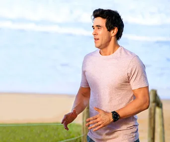 Man in a light pink t-shirt jogging on a beach path with ocean waves in the background.