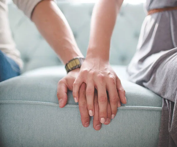 Two people sitting on a couch, gently holding hands, symbolizing connection and support.