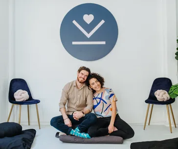 Couple sitting on cushions, smiling, with a large symbol on the wall behind them in a meditation setting.