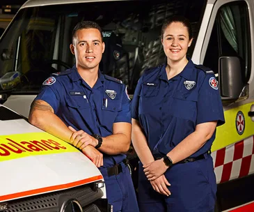 Paramedics in uniform standing in front of an ambulance, smiling confidently.