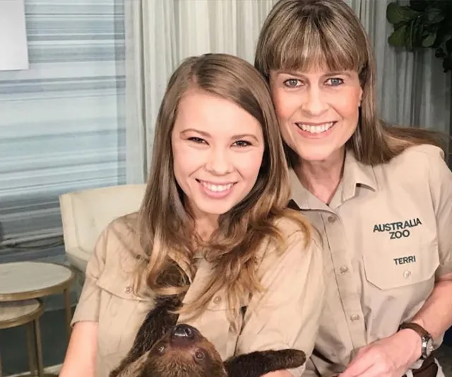 Two women smiling, one holding a small animal, both wearing khaki shirts with Australia Zoo logos indoors.