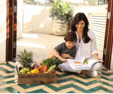Woman and child reading a book indoors, with a box of fresh fruits and vegetables beside them.