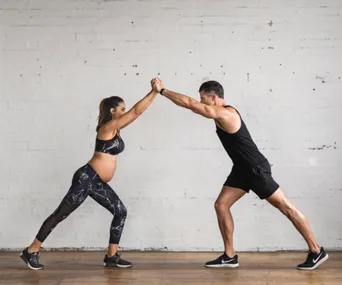 Pregnant woman and man performing partner exercise; hands clasped, both in athletic wear.