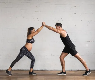Pregnant woman and man performing partner exercise; hands clasped, both in athletic wear.
