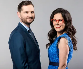 Two individuals in formal attire, a man in a suit and a woman in a blue dress with red glasses, posing and smiling.