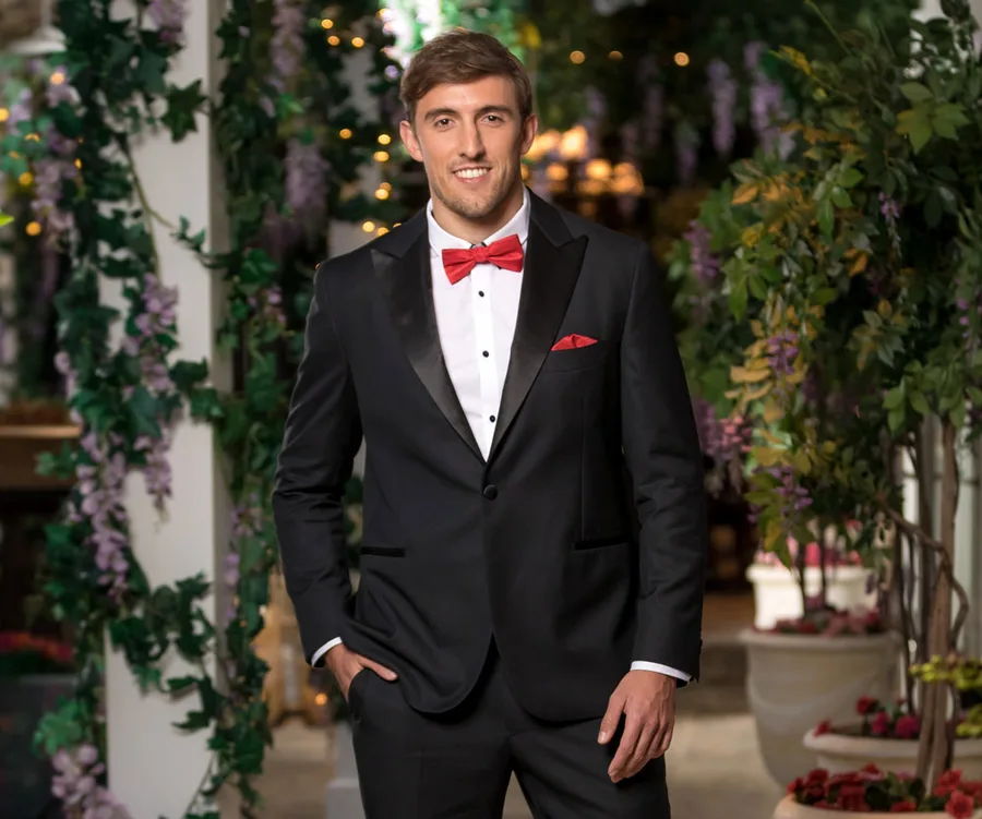 Man in a black tuxedo and red bow tie, standing in a garden setting with floral decorations.