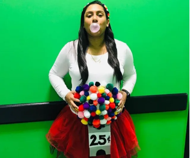 Woman dressed as a gumball machine, blowing bubblegum, with colorful pom-poms on her belly and 25ยข sign.