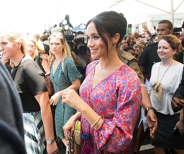 Meghan Markle in a colorful dress smiles while walking through a crowd in Fiji, surrounded by security and photographers.