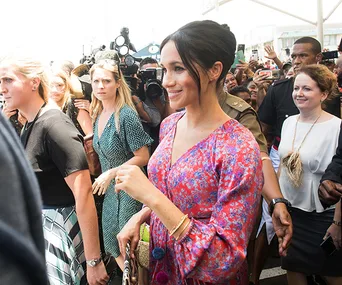 Meghan Markle in a colorful dress smiles while walking through a crowd in Fiji, surrounded by security and photographers.