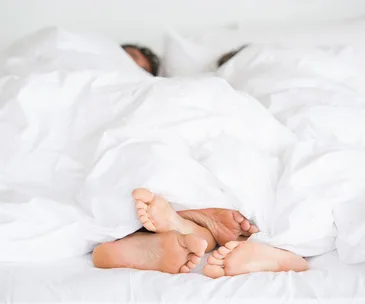 Two pairs of feet peeking out from under a white comforter, suggesting a couple lying in bed.