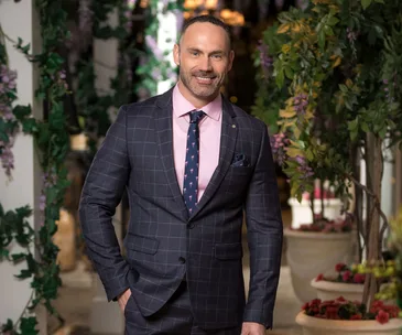 Man in a checked suit with a pink shirt and dark tie, standing in a garden setting with flowering plants.