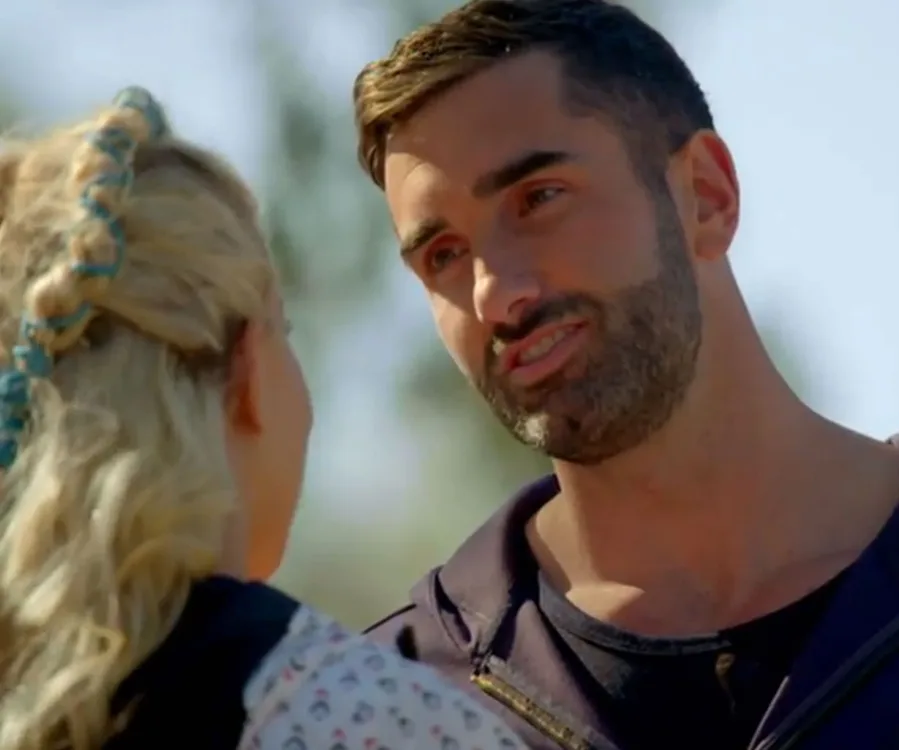 Man with short hair and beard talking to a woman with braided hair, outdoors under clear sky.