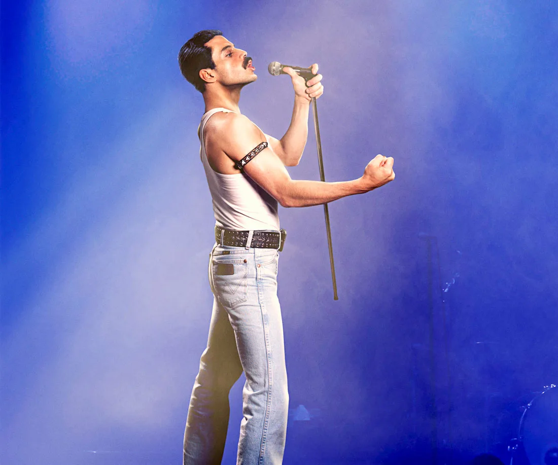 Actor in a white tank top and jeans sings on stage with a microphone stand, set in a blue-hued spotlight.