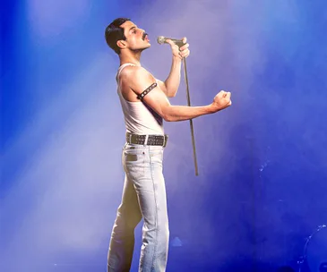 Actor in a white tank top and jeans sings on stage with a microphone stand, set in a blue-hued spotlight.