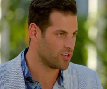 A man in a light striped suit and floral shirt, outdoors with blurred greenery background, engaged in conversation.