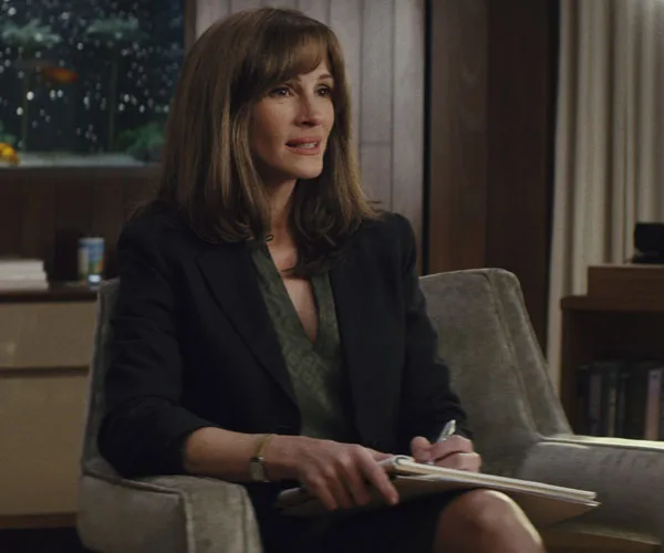 Julia Roberts in a dark blazer sits with notepad, pensively listening, in a scene from "Homecoming," an Amazon series.