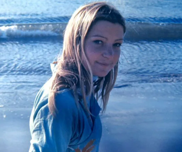 A woman with long hair in a blue top stands by the ocean, looking back at the camera.