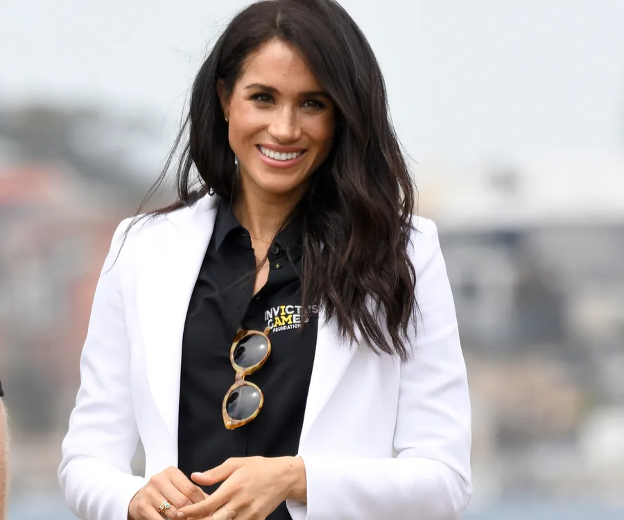 Smiling woman with long hair in a white blazer, holding sunglasses, outdoors.