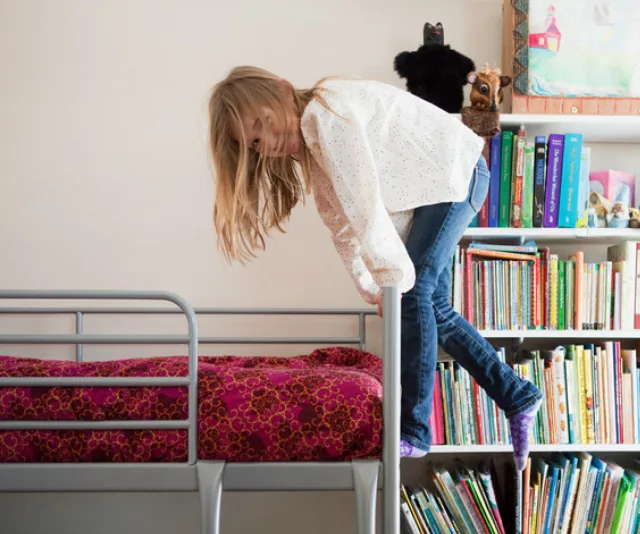 Child climbing a bunk bed ladder, surrounded by a bookshelf with colorful books and stuffed animals on top.