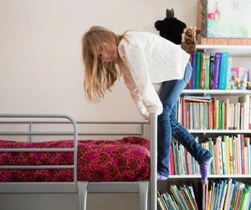 Child climbing a bunk bed ladder, surrounded by a bookshelf with colorful books and stuffed animals on top.