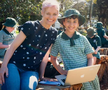 Smiling woman and child with a laptop outdoors, surrounded by other children in uniforms.