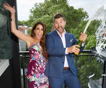 Man in suit popping champagne, woman in floral dress cheering, with trees in the background.