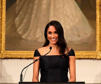 Meghan Markle giving a speech in New Zealand, standing at a podium, smiling in front of a framed painting.