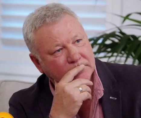 Man with gray hair and beard, wearing a black jacket and pink shirt, looking pensive while resting chin on hand indoors.