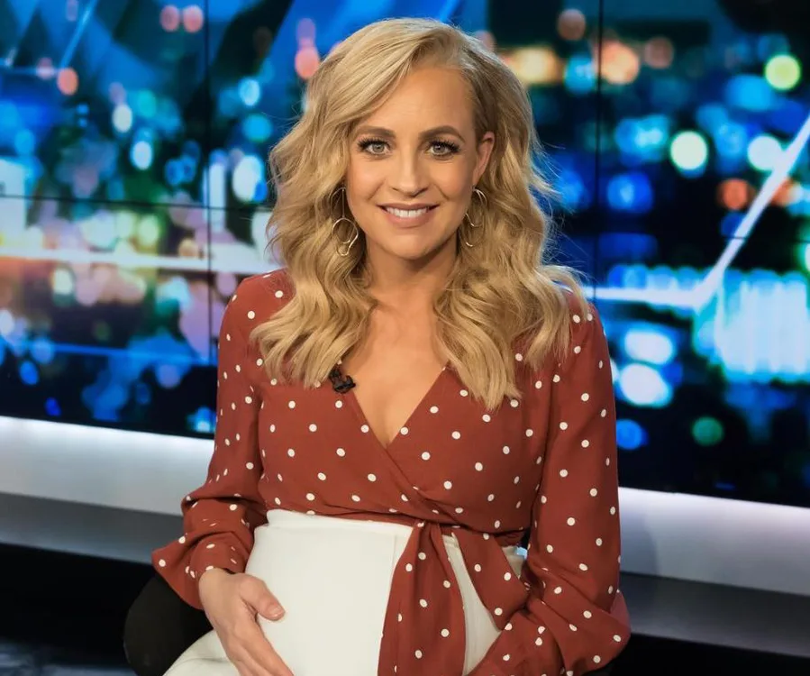 Blonde woman with long curls in a red polka-dot blouse, posing in a studio with a blurred city lights background.