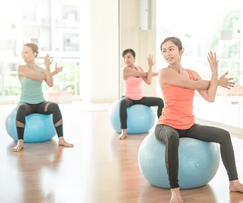 Women performing seated twists on exercise balls in a bright fitness studio.