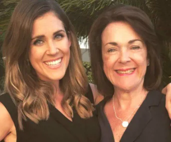 Two women smiling together outdoors, with green foliage in the background.