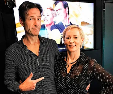 Two smiling individuals in front of a screen displaying a promotional image, one person pointing at the other.