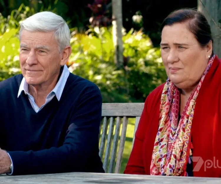 Two people sitting on a wooden bench outdoors, one in a navy sweater and the other in a red cardigan, with greenery behind.