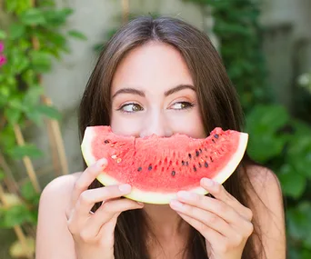 Woman playfully holding a watermelon slice in front of her face, looking to the side, outdoors in a garden.