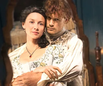 A couple embraces lovingly, reflected in a mirror, wearing period clothing from "Outlander" Season 4.