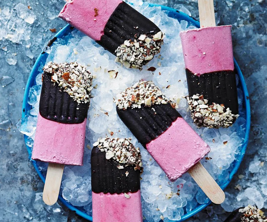 Pink and black vegan ice cream popsicles, coated with nuts, on a plate with ice.