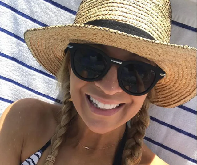 A person in a straw hat and sunglasses smiles, lying on a striped towel at the beach.
