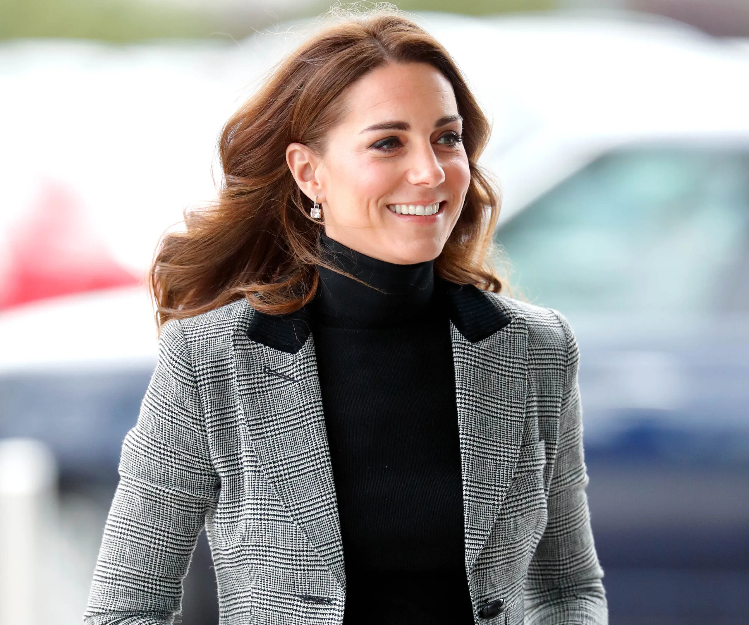 A woman with long brown hair smiles, wearing a black turtleneck and gray plaid blazer.