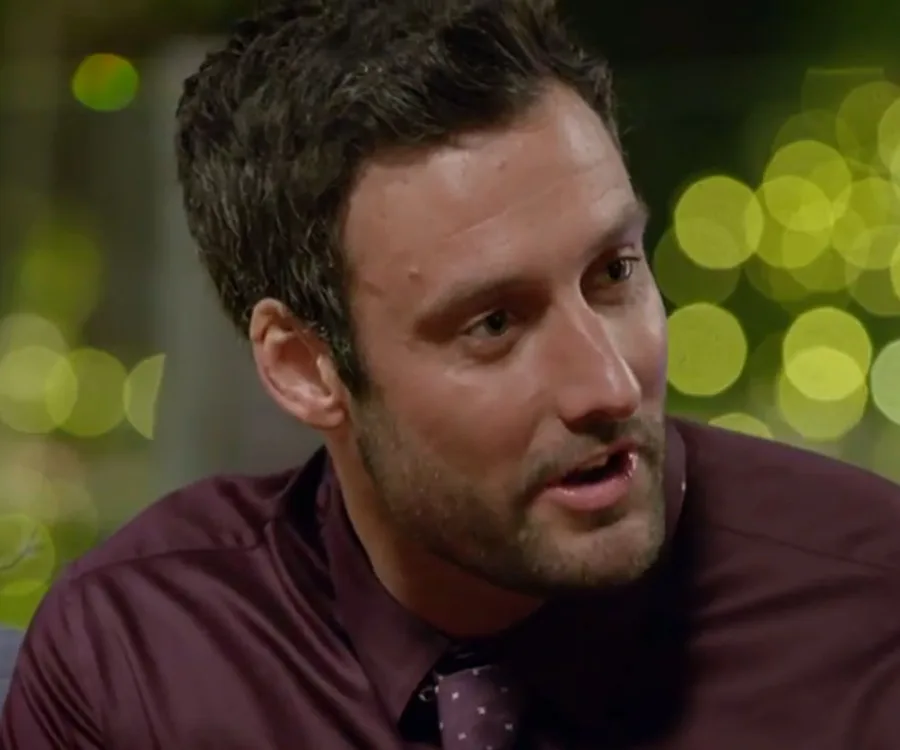 A man with dark hair in a maroon shirt speaks with blurred lights in the background.