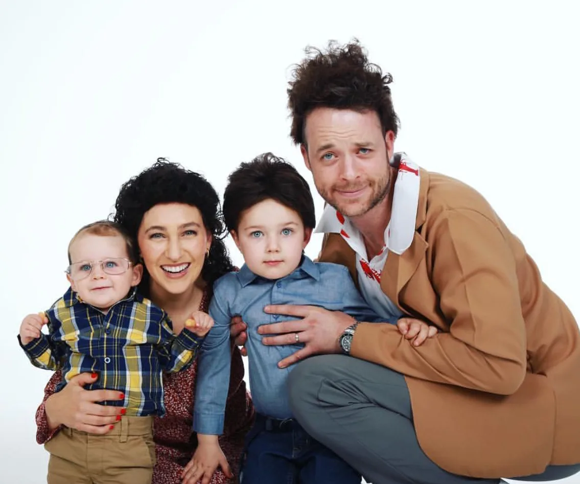 A family dressed in comedy characters' costumes, smiling together, with one adult in a brown jacket and two kids.