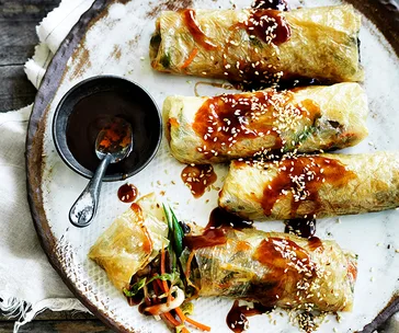 Crunchy vegan spring rolls drizzled with sauce and sprinkled with sesame seeds, served with dip on a rustic plate.