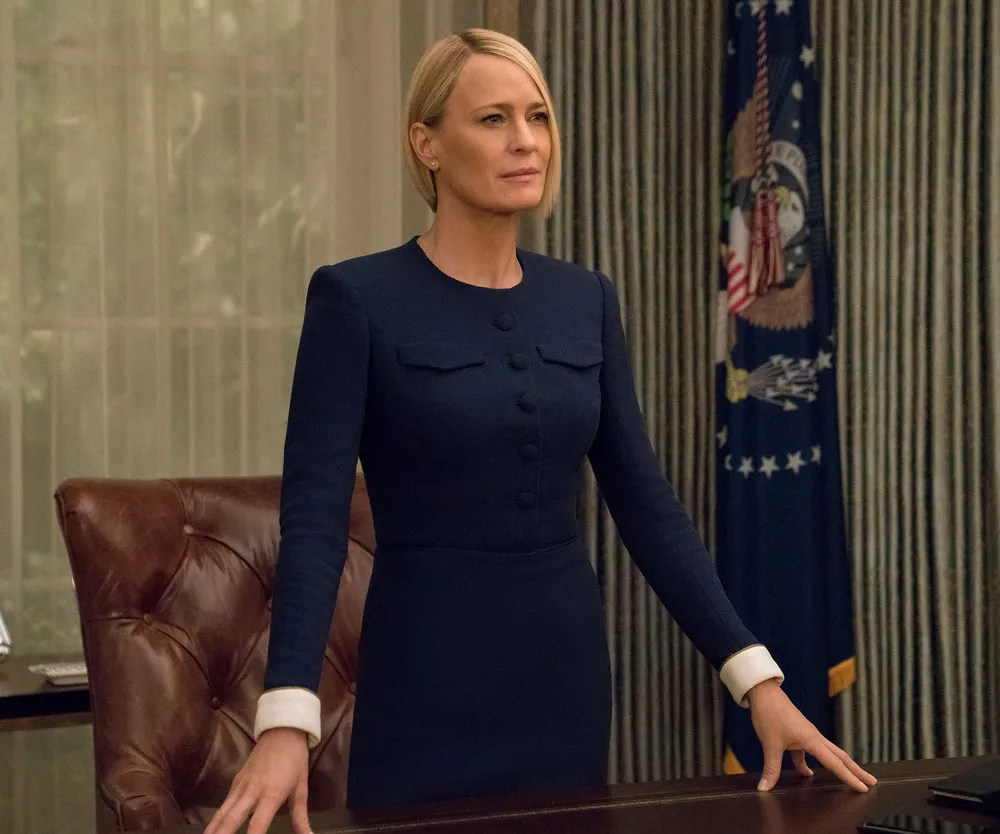 Claire Underwood stands confidently at a desk in a blue dress, with the U.S. flag behind her.