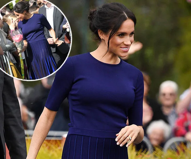 Meghan Markle wearing a navy dress, walking outside, greeting crowds with a smile.