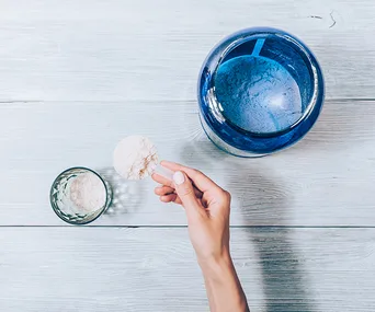 A hand scooping protein powder from a blue jar into a small glass on a light wooden surface.