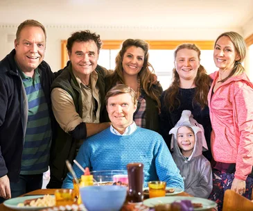 A group of seven people smiling at a table, with a young child in a dolphin costume in a bright room.