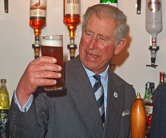 A man in a suit holds a pint of beer, inspecting it closely at a bar with bottles in the background.